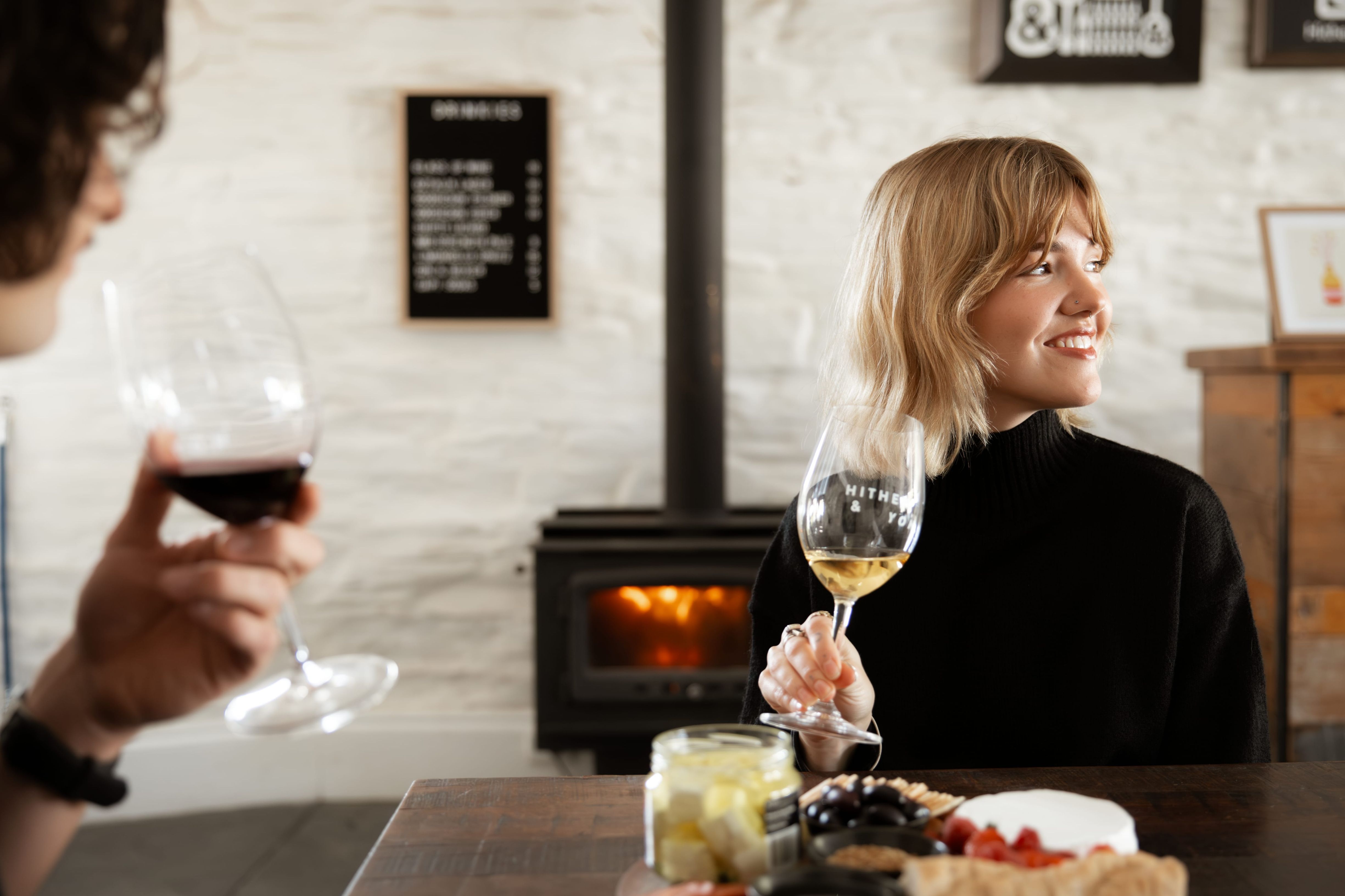 Lady in black top and blonde hair holding a glass of wine sitting in front of the fireplace at Hither and Yon