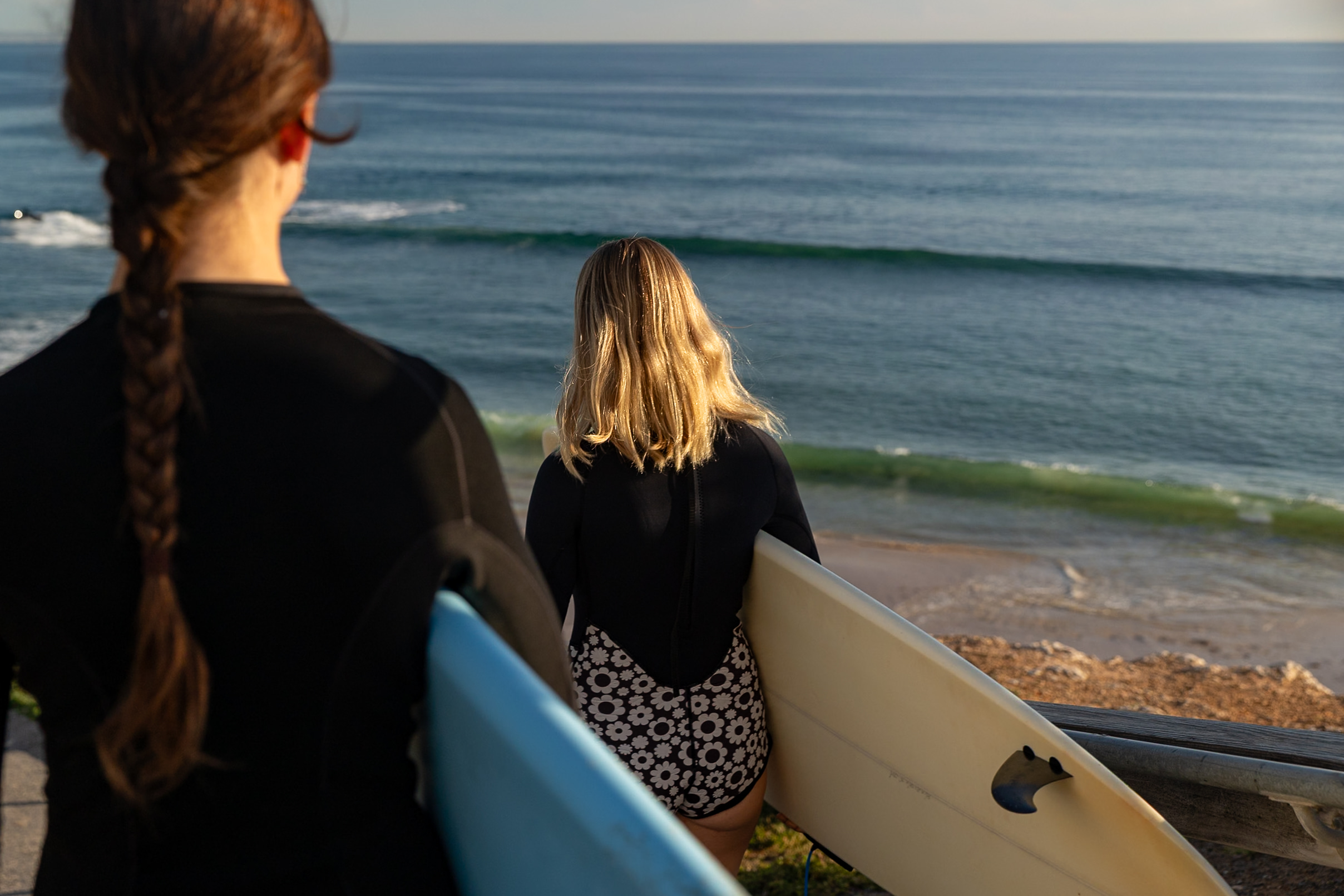 Surfers pausing to check the swell from the shoreline, boards in hand, golden hour light on the water